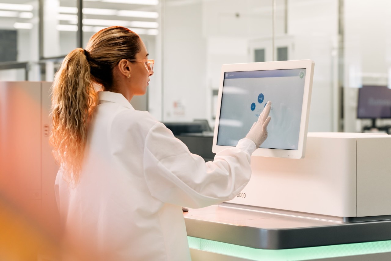 Close up, side view of a female scientist interacting with the touch screen monitor on a NovaSeq 6000, selecting from the sequencing screen; green status bar; blurry image in the foreground