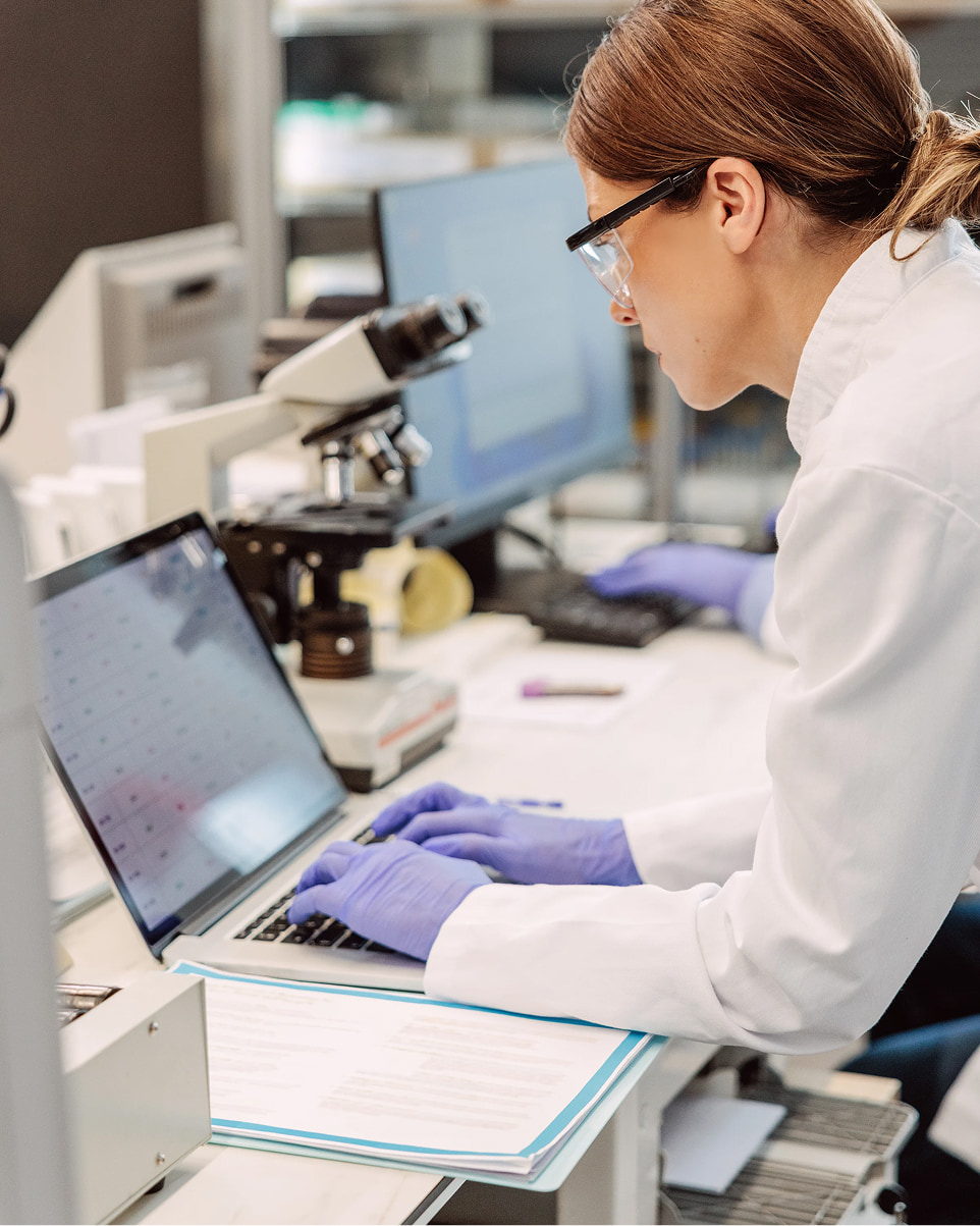 Profile view of a female scientist looking at a screen and typing on a keyboard of a laptop; located in a lab office with printed reports on the table, a microscope, another scientist, and other monitors blurry in the background.