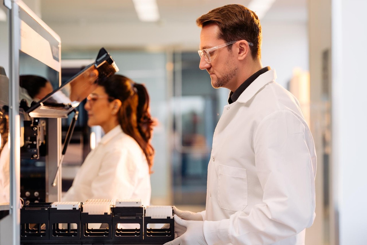 Profile image of a male scientist pushing the loading tray, filled with plates on a carrier of a Hamilton ML Star in preparation for a run in a lab setting; female scientist blurry in the background working