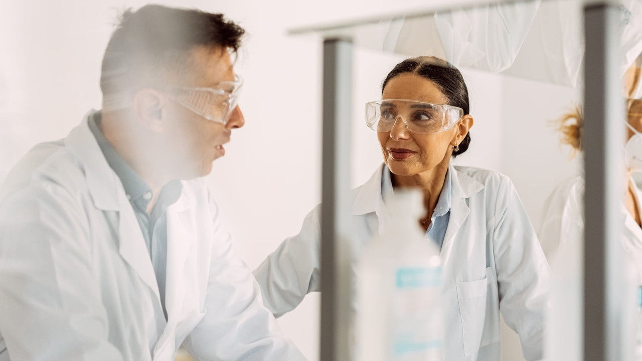  Two scientists, one male and one female, engaging in conversation in a lab; tubes and other lab equipment blurry in the foreground. 2025 Getty custom content