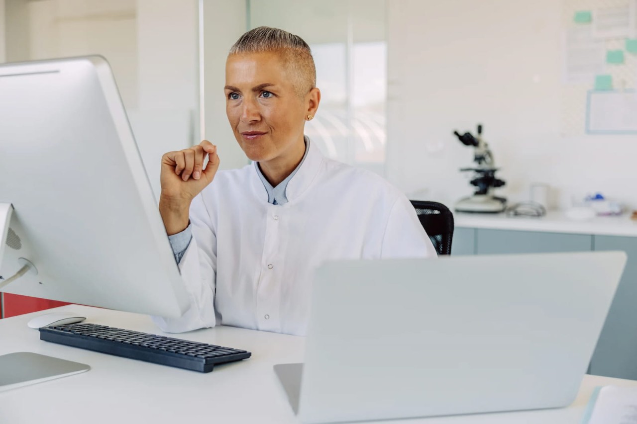 Front view of a female sitting at a desk in front of a desktop computer with an open laptop next to her; she is smiling and looking at the monitor; blurry in the background is a microscope and other papers and post-its pinned to a board.